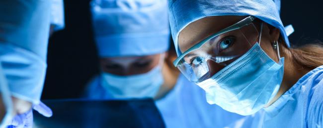 Close up of a doctor in scrubs, wearing safety glasses, looking at the camera with colleagues in the background