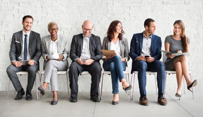 Group of 6 people in suits sitting in chairs waiting for an interview.